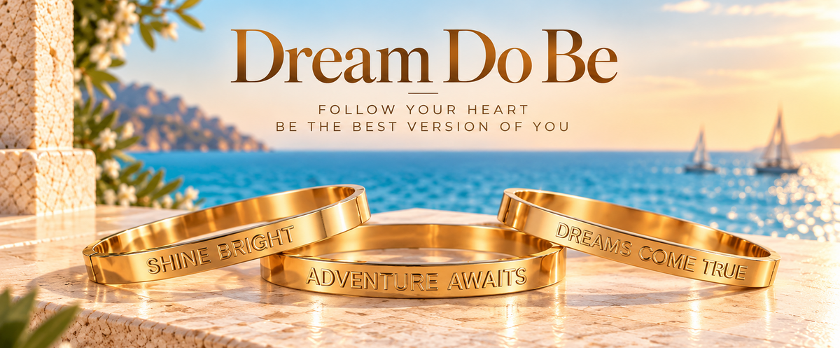 Three gold engraved bracelets displayed on a stone ledge with a sunny sea backdrop and the text 'Dream Do Be' above them.